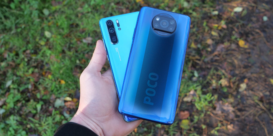 Hand holding two blue smartphones outdoors, one Xiaomi Poco with round camera and one Huawei with vertical cameras