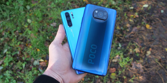 Hand holding two blue smartphones outdoors, one Xiaomi Poco with round camera and one Huawei with vertical cameras
