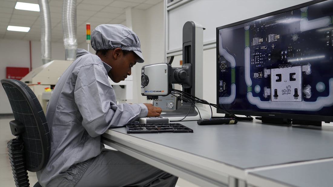 Technician in cleanroom outfit inspecting circuit board with EVO CAM microscope and large screen display