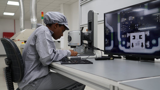Technician in cleanroom outfit inspecting circuit board with EVO CAM microscope and large screen display