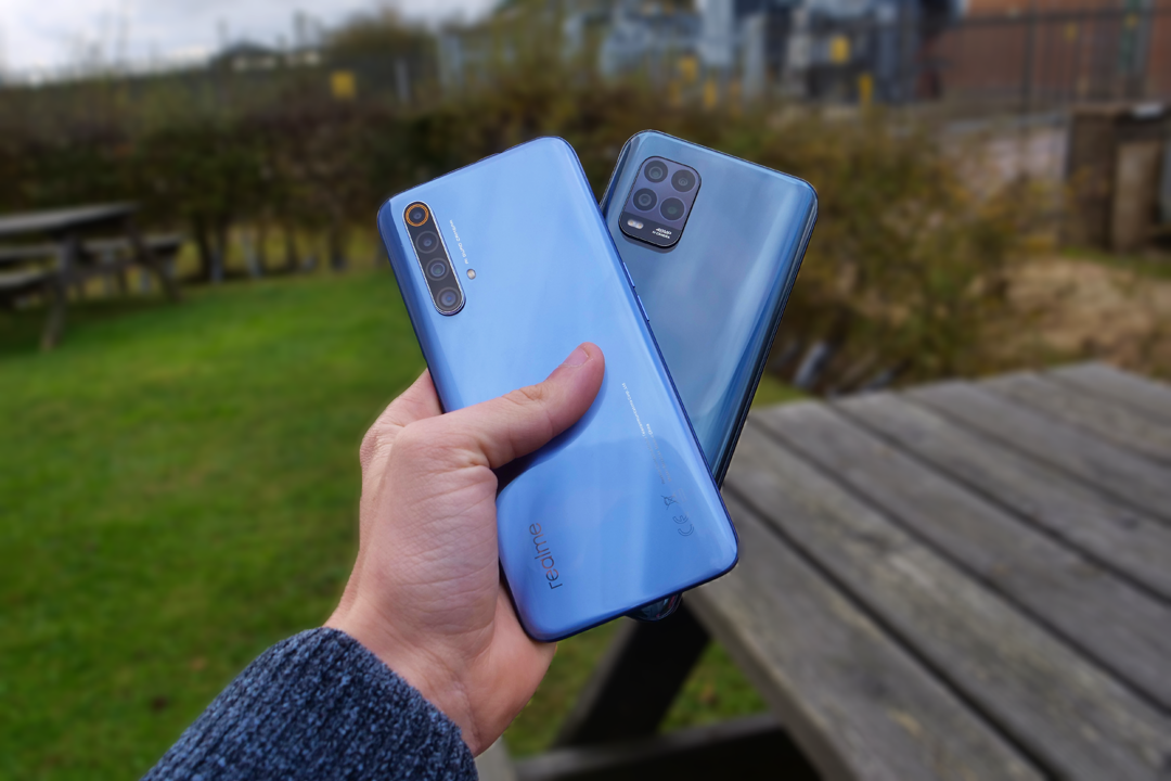 Person holding blue Realme and Xiaomi Mi 10 Lite 5G smartphones outdoors on a wooden table background
