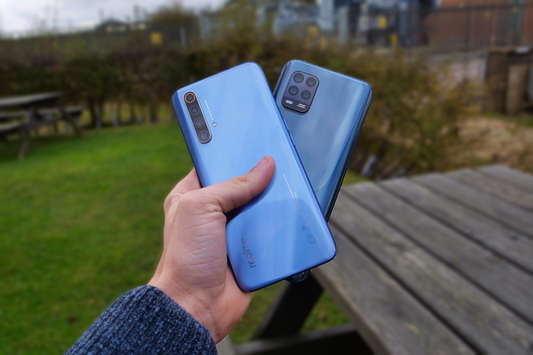 Person holding blue Realme and Xiaomi Mi 10 Lite 5G smartphones outdoors on a wooden table background