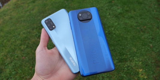 Hand holding blue Xiaomi POCO X3 NFC and light blue Realme 7 Pro smartphones against grass background