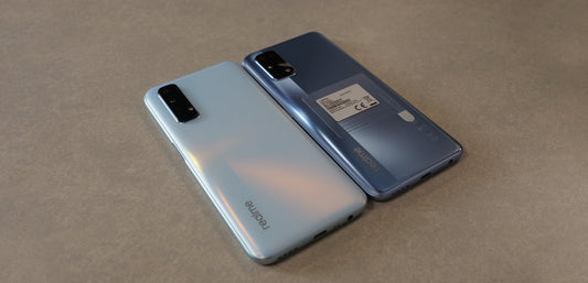 Two Realme smartphones with triple cameras on a grey surface, one silver and one dark blue, showcasing sleek design